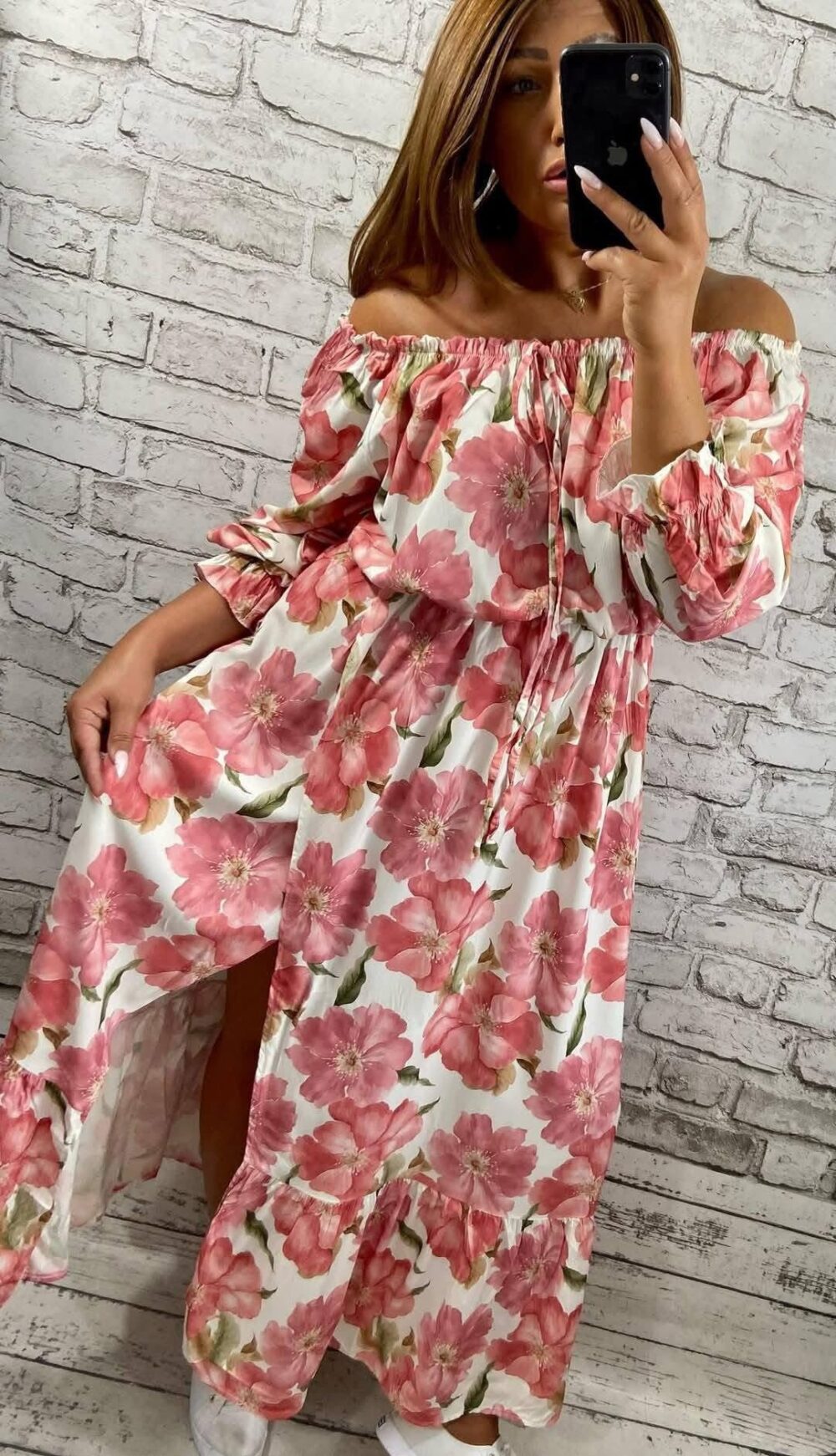 Žena v dlhých krémových šatách s ružovými kvetmi a odhalenými ramenami stojí pred svetlou tehlovou stenou