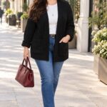 urob alt text k fotografii Plus size žena s dlhými hnedými vlasmi v čiernom pletenom kardigáne s vreckami, bielom tope a modrých džínsoch. Outfit dopĺňajú čierne členkové čižmy, bordová kabelka a zlaté náušnice. Jesenná prechádzka slnečným mestom.