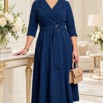 Plus size modelka v tmavomodrých midi šatách s prekladaným výstrihom, trojštvrťovými rukávmi, rozšírenou sukňou a opaskom so striebornou prackou. Outfit dopĺňa béžová kabelka a lodičky.