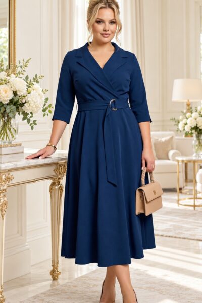 Plus size modelka v tmavomodrých midi šatách s prekladaným výstrihom, trojštvrťovými rukávmi, rozšírenou sukňou a opaskom so striebornou prackou. Outfit dopĺňa béžová kabelka a lodičky.