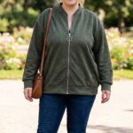 Plus size zelená bomber bunda so semišovým vzhľadom, zipsom, stojatým rebrovaným golierom, vreckami v švoch a elastickými lemami, kombinovaná s džínsami.