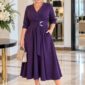 Plus size žena vo fialových elegantných šatách s kvetom v páse, béžovou kabelkou a lodičkami.