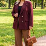 Plus size žena v bordovom predĺženom saku v dôstojníckom štýle s výraznými klopami, ozdobnými látkovými aplikáciami a reliéfnymi kovovými gombíkmi. Elegantné sako s voľnejším strihom a dekoratívnymi klopami imitujúcimi vrecká.