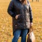 Plus size žena v čiernej mierne lesklej prešívanej bunde s kapucňou, predĺženým zadným dielom, elastickými manžetami a vreckami, v modrých rifliach a čiernych členkových čižmách v jesennom parku.