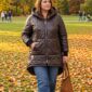 urob alt text Plus size žena v hnedej mierne lesklej prešívanej bunde s kapucňou, asymetrickou dĺžkou, predĺženým zadným dielom, vreckami a elastickými manžetami, v svetlomodrých rifliach a čiernych topánkach v jesennom parku.