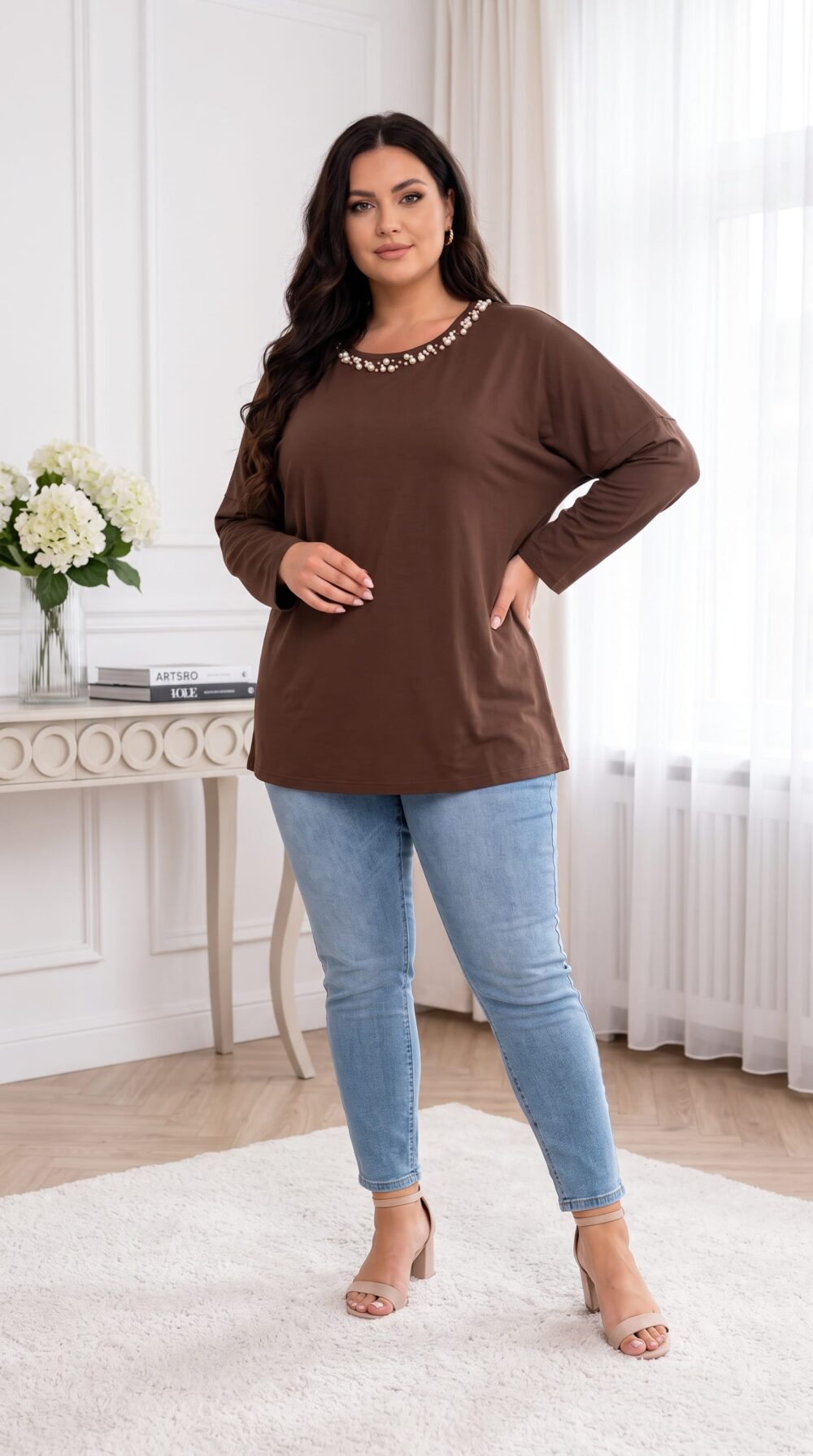 Plus size modelka v hnedej tunike s perlovým zdobením okolo výstrihu, spadnutými ramenami a dlhými rukávmi. Voľný, predĺžený strih dopĺňajú svetlomodré úzke rifle a béžové sandále na podpätku.