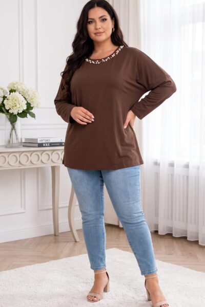 Plus size modelka v hnedej tunike s perlovým zdobením okolo výstrihu, spadnutými ramenami a dlhými rukávmi. Voľný, predĺžený strih dopĺňajú svetlomodré úzke rifle a béžové sandále na podpätku.