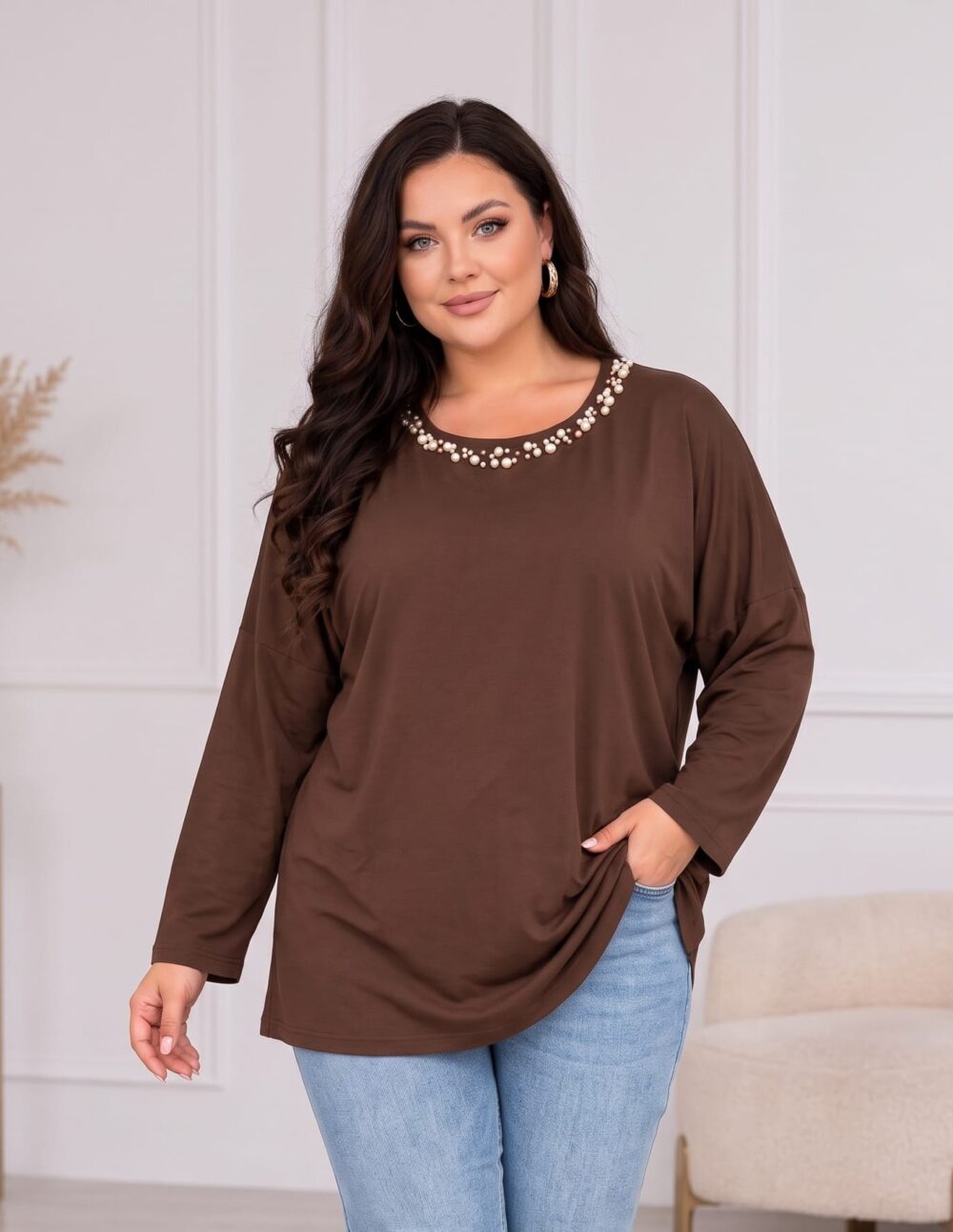 Hnedá dámska tunika vo voľnom strihu s dlhými rukávmi a okrúhlym výstrihom zdobeným bielymi perlami rôznych veľkostí. Plus size modelka ju kombinuje so svetlomodrými rifľami.