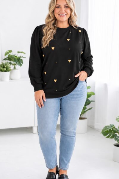 Plus size modelka v čiernej mikine so zlatými srdiečkami a drobnými zlatými ozdobami, v svetlomodrých džínsoch a čiernych mokasínach, v elegantnom svetlom interiéri.