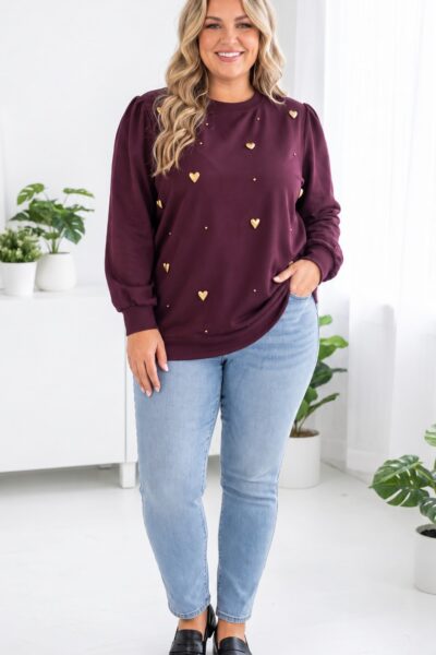 Usmievavá plus size žena vo fialovej mikine so zlatými plastickými srdiečkami a drobnými cvočkami, svetlomodrých džínsoch a čiernych mokasínach v svetlom elegantnom interiéri.
