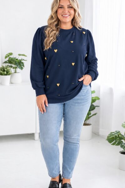 Plus size modelka v tmavomodrej mikine so zlatými plastickými srdiečkami a drobnými cvočkami, s jemne nariasenými rukávmi, svetlomodrými džínsami a čiernymi mokasínami v modernom bielom interiéri.