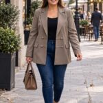 Plus size žena v predĺženom hnedobéžovom saku s ozdobnými striebornými gombíkmi a látkovými aplikáciami, tmavomodrých džínsoch a koňakových lodičkách kráča ulicou európskeho mesta s kabelkou v ruke.