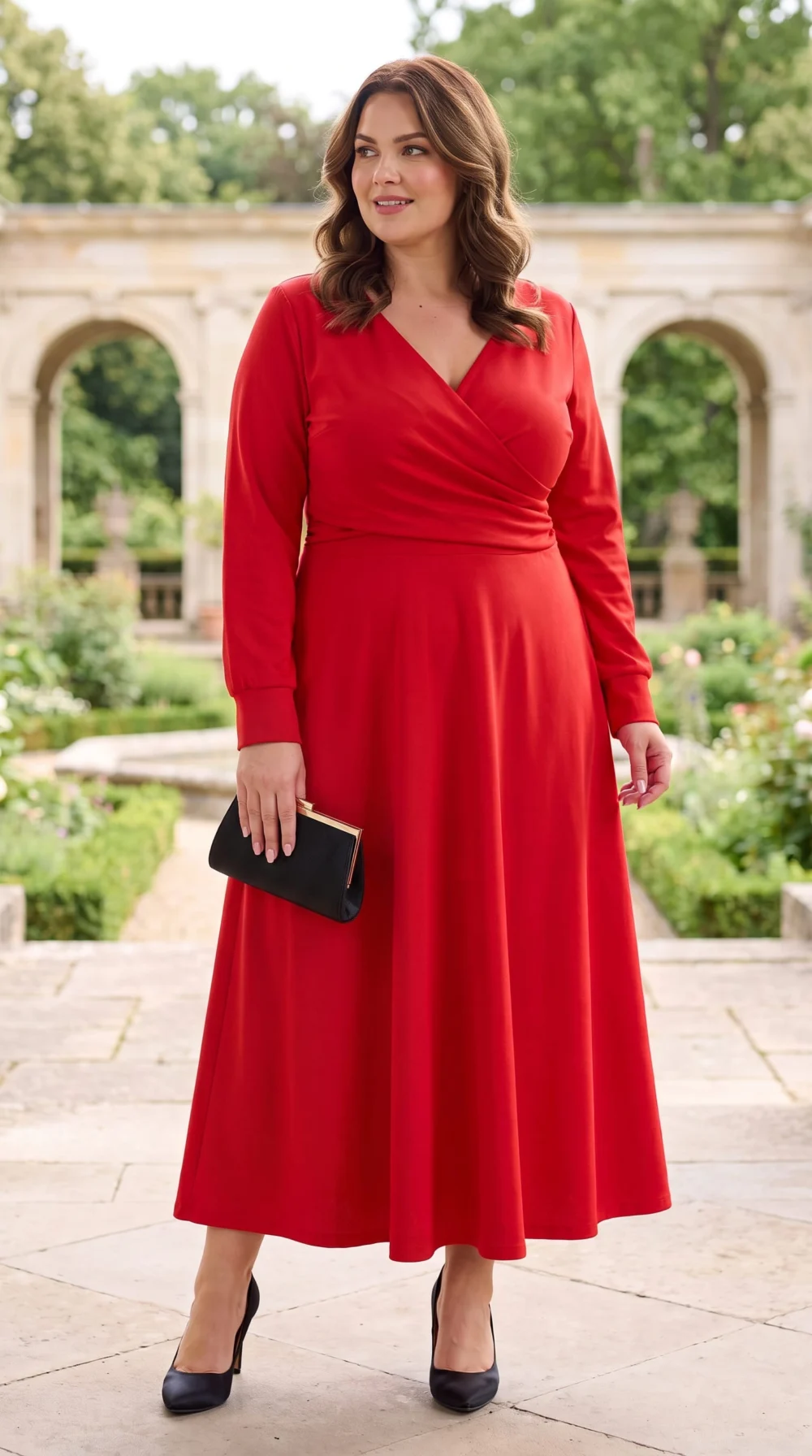 Elegantné červené dámske maxi šaty pre moletky s prekladaným výstrihom do V, jemným riasením v páse, dlhými rukávmi a rozšírenou sukňou. Plus size modelka s čiernou listovou kabelkou a lodičkami v romantickej záhrade.