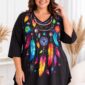 Plus size žena v čiernej tunike s výrazným farebným vzorom pierok, kvetov a lapača snov. Tunika má V-výstrih, trojštvrťové rukávy a voľný strih s predĺženými bokmi. Modelka pózuje v svetlom elegantnom interiéri.