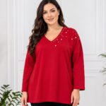 Detail červenej plus size tuniky s voľným strihom, dlhými rukávmi a V-výstrihom. Biele perly rôznych veľkostí zdobia asymetricky jednu stranu výstrihu, rameno a hornú časť tuniky.