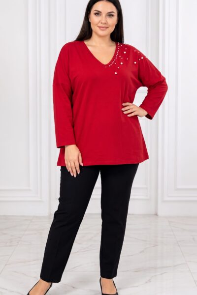 Plus size modelka v červenej tunike voľného strihu s V-výstrihom, dlhými rukávmi a asymetrickým perlovým zdobením na ramene a okolo výstrihu. Tuniku kombinuje s čiernymi nohavicami a lodičkami, jednu ruku má vbok.