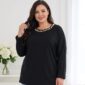 Plus size žena v čiernej tunike voľného strihu s dlhými rukávmi a okrúhlym výstrihom zdobeným bielymi perličkami. Detail tuniky v kombinácii so svetlomodrými rifľami.