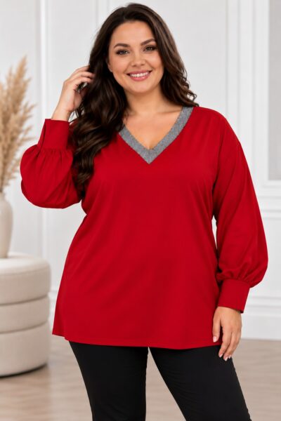 Plus size modelka v červenej tunike s voľným strihom, striebristosivým lemovaním V-výstrihu a dlhými balónovými rukávmi zakončenými manžetami. Tuniku dopĺňajú čierne nohavice. Modelka stojí vo svetlom elegantnom interiéri.